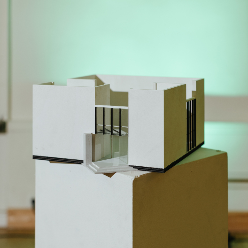 ID: Een maquette van het Rietveldpaviljoen op een witte sokkel. Het Rietveldgebouw is ontworpen door Gerrit Rietveld in 1954, het is een vierkant modernistisch gebouw met witte muren.