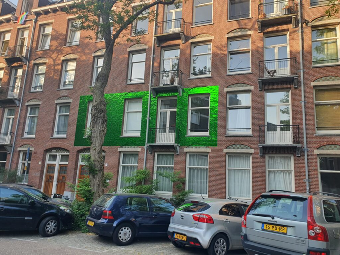 op de gevel van de tweede verdieping van een rijtjeshuis is het kunstwerk van Esther Tielemans te zien. Dit kunstwerk bestaat uit een glanzend groen vlak dat 3 ramen en 1 balkon omringt. Voor de rijtjeshuizen staat een boom en er is een parkeerplaats.